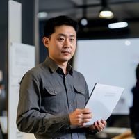 蘑菇视频官网用户体验负责人陈龙的专业形象照，男性，着浅色衬衫，手持平板设备展示产品界面，背景为用户研究白板，氛围开放创新